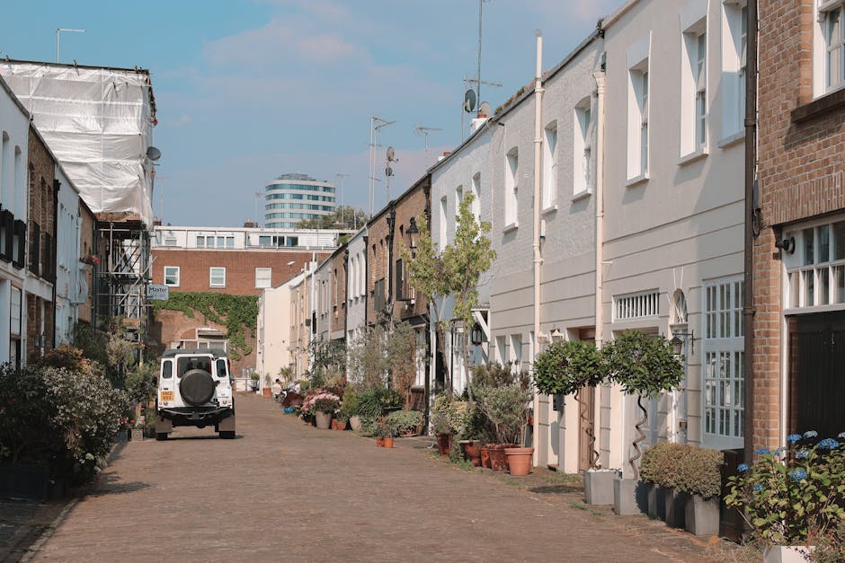 A narrow, cobblestone courtyard flanked by white residential buildings featuring large windows with black railings and flower boxes. Potted plants, including small trees and shrubs, line both sides of