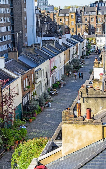 A narrow cobblestone street lined with small, uniform white and beige terraced houses that feature pitched slate roofs and small front gardens filled with potted plants and shrubs. The street is locat